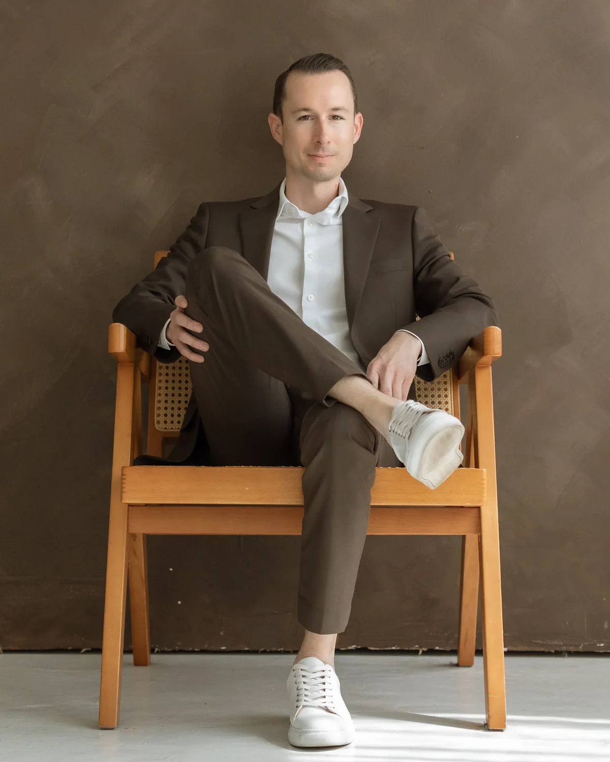 Justin Taylor seated in a wooden chair, wearing a brown suit against a warm textured wall